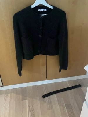 Svart/ mörkgrå stickad kofta från Zara - Mysig svart stickad kofta från Zara med rund hals, stora framfickor och knappar framtill. Koftan har lång ärm och en croppad passform. Perfekt till lager på lager under höst och vinter.