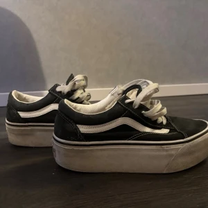 Svarta Vans sneakers med platåsula - Säljer ett par klassiska Vans sneakers i svart och vitt med platåsula. Skorna har vit snörning, rund tå och den ikoniska vågiga Vans-detaljen på sidan. Ovandelen är i canvas och skinn, och sulan är tjock och vit för extra höjd.