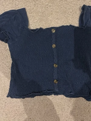 Blå croppad blus med knappar - Säljer en blå croppad blus med korta puffärmar och smockad struktur. Toppen har dekorativa knappar framtill och en lätt vågig kant nertill. Toppen är i storlek 140, men motsvarar en S!