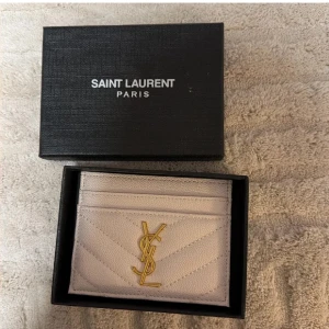 Vit Saint Laurent kortplånbok i skinn - Supersnygg vit kortplånbok från Saint Laurent med guldigt YSL-logo framtill. Plånboken är gjord i lyxigt skinn med quiltad struktur och har flera kortfack både fram och bak. Perfekt för dig som vill ha något stilrent och exklusivt i väskan.