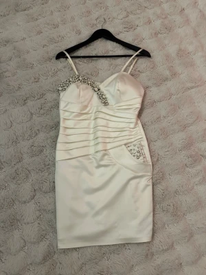 Vit kort klänning med pärlor och strass -  Elegant kort klänning av satin med strassdetaljer runt halsringningen och på fickan. Klänningen har smala axelband och en figurnära passform med rynkade detaljer framtill. Den stängs med dragkedja baktill och har en liten sprund nedtill. Några stenar har trillat av, små lösa trådar förekommer se bilder. Har använt den två gånger och har inte testat att tvätta den. Perfekt till studenten eller ditt bröllop. Upplevs mer som 36, crèmefärgad