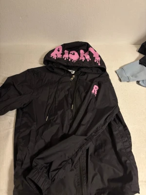 Svart vindjacka från Risky XS - Svart vindjacka från Risky i storlek XS. Jackan har huva med stora rosa bokstäver och dragkedja framtill. Tillverkad i lätt polyester med justerbar dragsko i huvan och fickor på sidorna. Perfekt för dig som gillar streetwear och vill sticka ut.