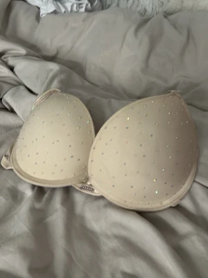 Beige bh med glittriga detaljer - Snygg beige bh med vadderade kupor och glittriga paljettdetaljer över hela framsidan och axelbanden. Bh:n har en enkel och stilren design med justerbara axelband och hakknäppning bak. Perfekt för dig som vill ha något extra under kläderna.