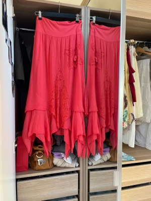 Rosa långkjol med spetsdetaljer - Korallrosa långkjol med asymmetrisk, vågig fåll och spetsinslag framtill. Kjolen har bred midja och dekorativa spetsapplikationer. Från vintage butiken CRUSH i Köpenhamn för 350 DKK (≈500kr).  Fint skick! Öppen för bud (vänligen lägg endast bud vid seriöst köpintresse).