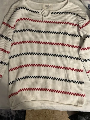 Randig stickad tröja från Marine Sweden - Säljer en vit stickad tröja från Marine Sweden i storlek S/36. Tröjan har ribbad struktur, rund halsringning med snörning och horisontella ränder i svart och rött. Perfekt för dig som gillar klassisk men ändå chill stil.