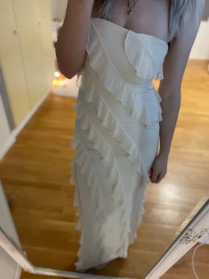 Vit axelbandslös långklänning med volanger - Säljer en superfin vit långklänning med axelbandslös design och diagonala volanger över hela klänningen. Klänningen har en elegant och figurnära passform som ger en snygg siluett. Perfekt för dig som vill sticka ut med en romantisk look.