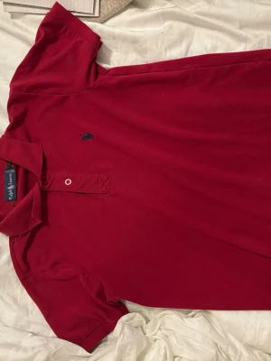 Vinröd pikétröja från Ralph Lauren - Klassisk vinröd röd pikétröja från Ralph Lauren med blå broderad logga på bröstet. Tröjan har krage, två knappar framtill och korta ärmar. Perfekt skick!