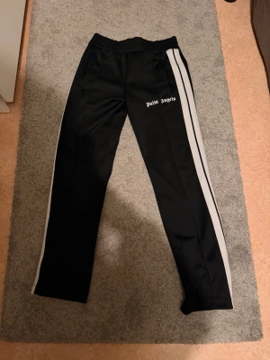 Palm Angels svarta trackpants  - Säljer ett par svarta trackpants från Palm Angels i storlek S. Byxorna har vita dubbla sidoremsor, elastisk midja och logga broderad på vänster sida. Tillverkade i 100% polyester för en sportig vibe och skön passform. Har alla lappar och äkthetsbevis ,bättre bilder går att fås vid intresse