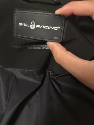Svart Sail Racing dunjacka med huva - Svart dunjacka från Sail Racing med huva och dragkedja vill inte sälja vill ha byte med samma jacka fast i storlek 150 och möts i göteborg