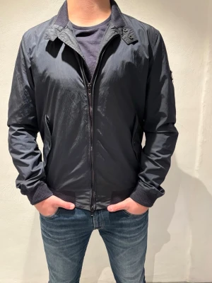 J.Lindeberg jacka - J.Lindeberg jacka i mörkblå färg, ståkrage och dragkedja framtill. Jackan har diskret logga på ärmen och två fickor. Perfekt till jeans och t-shirt för en clean look. Storleken är i M