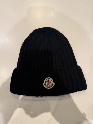 Svart mössa/beanie från Moncler - Säljer en svart ribbstickad mössa från Moncler med klassisk logga framtill. Mössan är i mjuk ull och har vikt kant för extra stilpoäng. Perfekt accessoar för kalla dagar och snygg till alla outfits.
