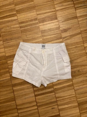 Vita cargo shorts från BDG Urban Outfitters - Säljer ett par vita cargo shorts från BDG Urban Outfitters i storlek XS. De har elastisk midja, stora fickor på sidorna och bak samt knapp och dragkedja framtill. Perfekta för varma dagar och chill stil.