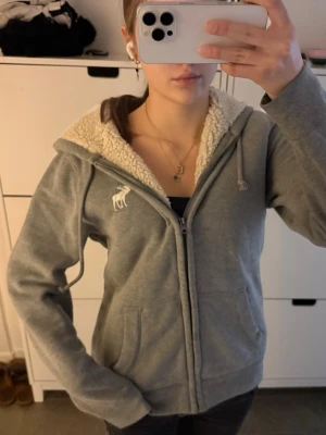 Fodrad zip up hoodie Abercrombie and fitch  - Storlek S! ❤️ Mysig grå hoodie från Abercrombie & Fitch med dragkedja, vit broderad älg på bröstet och fluffigt fuskpälsfoder i huvan. Tröjan har snörning i huvan och två fickor framtill. Perfekt för kyliga dagar.