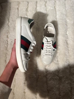 Vita Gucci ace - Exklusiva Gucci sneakers i äkta läder med klassiska gröna/röda ränder och präglad Gucci-text baktill. En tidlös och eftertraktad modell som fungerar perfekt till både vardag och uppklädda outfits.  ✔️ Märke: Gucci✔️ Storlek: EU 41,5✔️ Färg: Vit med gröna/röda detaljer✔️ Skick: Mycket bra begagnat skick✔️ Anmärkning: Endast lätt smuts på sulan (se bilder)  Skorna är väl omhändertagna, inga sprickor eller skador, och har mycket livslängd kvar. Perfekt tillfälle att köpa snygga Gucci ace till bra pris.