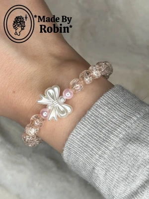 Armband ”Nina” 🎀🤍 - Vårt handgjorda armband ”Nina”  med transparenta pärlor och en vit rosett i mitten.🥰✨ Två ljusrosa pärlor sitter på varsin sida om rosetten och ger en gullig touch. Perfekt accessoar för dig som gillar detaljer och pastellfärger.