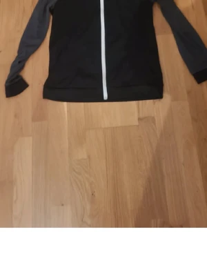 Svart och grå hoodie med dragkedja - Säljer en svart hoodie med grå ärmar och vit dragkedja framtill. Tröjan har lång ärm och är perfekt för dig som gillar en enkel och sportig stil. Passar lika bra till jeans som till träningsbyxor.