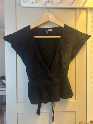 Svart omlottopp från Divided S - Säljer en svart omlottopp från Divided i storlek S. Toppen har en djup v-ringning, breda axlar och knytband i midjan som ger en snygg siluett. Tillverkad i ett lätt och luftigt tyg, perfekt för varmare dagar.