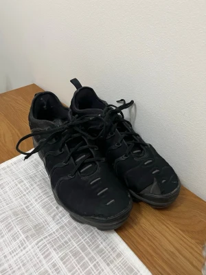 Svarta Nike sneakers med snörning - Säljer ett par helsvarta Nike sneakers med cool vågig design på sidan och klassisk snörning. Skorna har en chunky sula och diskret Nike-logga på sidan. Materialet är en mix av syntet och textil, perfekt för dig som gillar sportig streetstyle.