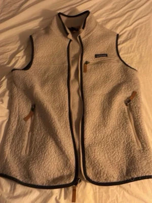 Beige fleeceväst från Patagonia - Säljer en beige fleeceväst från Patagonia i storlek S. Västen har dragkedja framtill, tre fickor med dragkedja och snygga kontrastkanter i svart. Perfekt för lager-på-lager och outdoor-vibe. Klassisk Patagonia-logga på bröstet och mjukt, mysigt material.