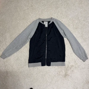 Blå bomberjacka från Made In The Shade - Snygg bomberjacka med marinblå kropp och ljusgrå ärmar från Made In The Shade. Jackan har dragkedja framtill, ribbade muddar och krage samt en clean, sportig look. Perfekt för dig som gillar en avslappnad men ändå stilren vibe.