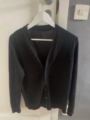 Svart merinoullskofta - Svart kofta i mjuk merinoullsblandning. Klassisk modell med v-ringning, knappar framtill och långa ärmar. Perfekt att slänga över en t-shirt för en clean och stilren look.