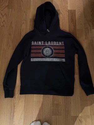 Saint Laurent hoodie S🤩 - Säljer en fet sällsynt ysl hoodie som har ett stort snyggt tryck på framsidan av hoodien  får en archive grisch stil, hoodien är väldigt enkel att styla och passar till olika färger. Storlek är S och passar 165-174 beroende på vikt.
