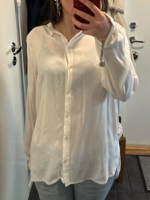 Vit skjorta i silkeslook från Lindex - Säljer en vit skjorta från Lindex i storlek M. Skjortan har en klassisk krage, knappar framtill, lång ärm och längre baktill. Materialet har en silkeslen känsla och ger ett snyggt, avslappnat intryck. 