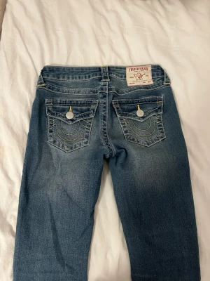 True Religion blå jeans med kontrastsömmar - Säljer ett par blå jeans från True Religion med ikoniska kontrastsömmar och snygga fickdetaljer bak. Jeansen har normal passform, klassisk femficksdesign och guldfärgade sömmar som sticker ut. Materialet är slitstarkt jeans och modellen har en rak siluett.