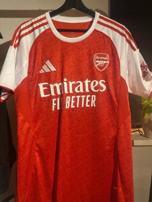 Arsenal Adidas matchtröja röd/vit M - Säljer en officiell Arsenal matchtröja från Adidas i rött och vitt med klubbmärke och Emirates-tryck på bröstet. Tröjan har ett subtilt mönster, vita axelpartier med röda ränder och är i slim fit. Perfekt för dig som älskar fotboll och Arsenal.