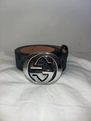 Svart GG bälte med silver spänne - Jag säljer ett helt nytt svart läderbälte med GG mönster och silver spänne. Detta bälte har dessutom ett gummi band för att inte tappa bort delen som ska hålla fast så att det inte fladdrar runder.