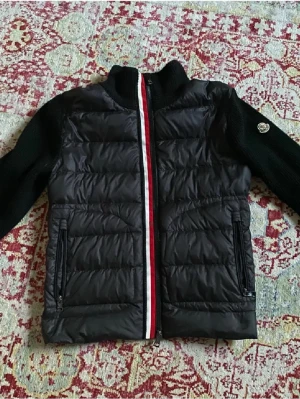 Svart Moncler pufferjacka med stickade ärmar - Säljer en svart cardiganjacka från Moncler med stickade långärmade ärmar och quiltad kropp. Jackan har dragkedja framtill med röd och vit rand, samt två fickor med dragkedja. Klassisk Moncler-logga på ena ärmen. Perfekt för höst och vinter.