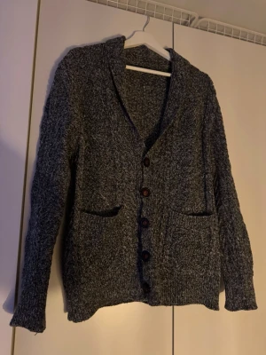 Mörkgrå stickad kofta med knappar - Säljer en mörkgrå stickad kofta från bellas Sverige med grovt mönster och stora bruna knappar. Koftan har två fickor framtill och lång ärm. Perfekt för kyliga dagar och passar till jeans eller över en t-shirt. Denna sitter dessutom perfekt och är väldigt stilren till alla stilar.