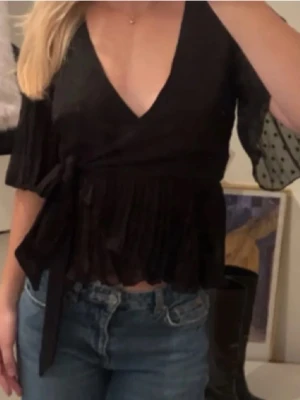 Svart omlottopp med knyt och volang - Säljer en svart omlottopp med knyt i midjan och volangkant nedtill. Toppen har korta, lätt transparenta ärmar med diskreta prickar och v-ringad urringning. Perfekt för dig som gillar trendiga detaljer och en feminin look.