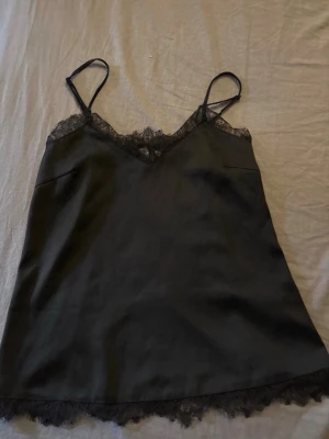 Svart linnetopp med spetsdetaljer - Säljer en svart linnetopp med tunna axelband och vacker spets längs urringningen och nederkanten. Toppen har en lätt glansig finish och känns riktigt mjuk och skön mot huden. Perfekt att styla lager på lager eller bära som den är.