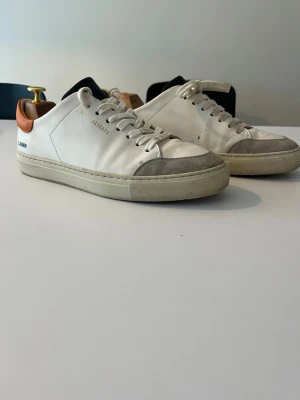 Vit sneaker från Axel Arigato med orange detalj - Snygga vita sneakers från Axel Arigato med orange häl och grå mockadetalj vid tån. Skorna har snörning, platt sula och diskret branding på sidan. Tillverkade i skinn och mocka, med en stilren och minimalistisk design som passar till det mesta.