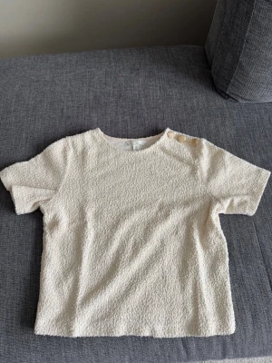 Krämvit topp/t-shirt med textur och knappar - Säljer en krämvit topp/t-shirt med korta ärmar och rund hals. Materialet är frottéliknande och ger en unik textur. På ena axeln finns tre dekorativa guldknappar som snygg detalj. Prislappen togs bort men tröjan kom aldrig till användning.