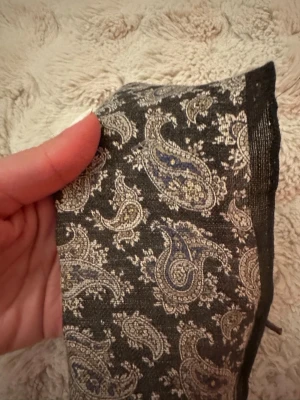 Svart paisleymönstrad näsduk Atlas Design - Säljer en svart näsduk mått 30x30  cm från Atlas Design med paisleymönster i beige och blå toner. Västen är tillverkad i 61% linne och 39% bomull, vilket ger en lätt och luftig känsla. Perfekt för dig som vill ha en unik och stilren look. Nypris 299 kr, aldrig använd