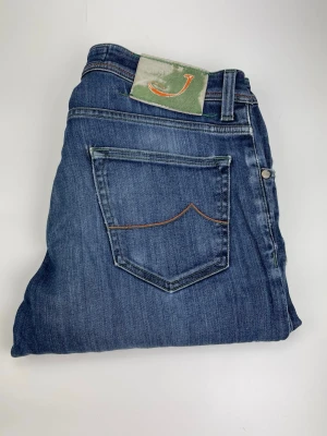 Jacob Cohen 622 - Snygga blå jeans från Jacob Cohen, modell Style 622. Klassisk femficksdesign med orange kontrastsömmar och knappgylf. Jeansen har en grön patch med broderad J på bakfickan och är tillverkade i mjukt denimtyg för en bekväm känsla.