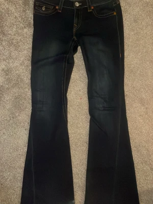 Mörkblå bootcut jeans True Religion - Säljer ett par mörkblå jeans från True Religion med kontrastsömmar och snygga fickdetaljer bak. Modellen har bootcut-ben och klassisk femficksdesign. Jeansen har silverfärgade knappar och nitar samt orangea sömmar som sticker ut. Materialet är robust jeans.