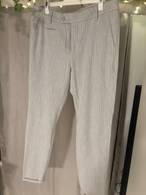 Grå kostymbyxor från Les Deux - Snygga grå kostymbyxor från Les Deux med tunna vita ränder. Byxorna har en klassisk rak passform, bälteshällor och knappstängning. Perfekta för en clean och stilren look. Materialet känns mjukt och har en lätt struktur.