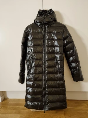 Svart lång dunjacka från Moncler - Säljer en svart, glansig lång dunjacka från Moncler med huva och dragkedja framtill. Jackan har Moncler-logga på ärmen, två fickor med dragkedja och quiltad design. Perfekt för kalla dagar när du vill vara både varm och snygg.