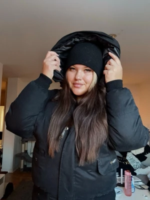 Svart pufferjacka från Canada Goose - Säljer en svart pufferjacka från Canada Goose med klassisk logga på ärmen och stor huva. Jackan har dragkedja framtill, stora fickor och är fodrad med dun för extra värme. Yttermaterialet är en mix av polyester och bomull, fodret är nylon. Perfekt för kalla dagar.