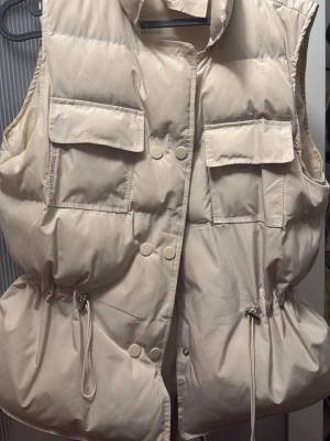 Beige dunväst med snörning - Säljer en beige dunväst med stora fickor framtill och snygga tryckknappar. Västen har dragsko i midjan för justerbar passform och en hög krage som ger extra stilpoäng. Perfekt att slänga över en hoodie eller tröja för en cool lager-på-lager-look.