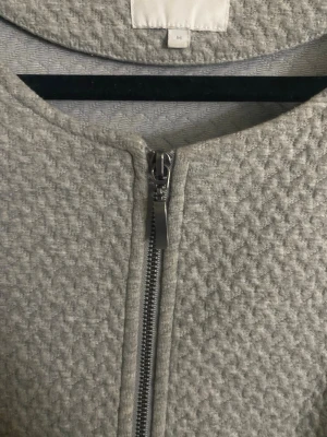 VILA Grey Textured Zip Jacket  - Stylish light grey jacket from VILA Clothes with a beautiful textured fabric. Features a minimalist round neckline and a full front zipper. Cropped length with long sleeves – perfect for both casual and smart outfits.  Easy to style with jeans, trousers, skirts, or dresses. Great as a lightweight jacket for spring, autumn, or indoor wear.