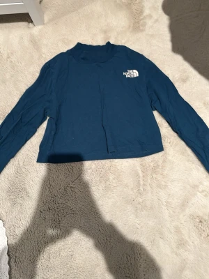Blå långärmad topp från The North Face - Säljer en blå långärmad topp från The North Face med liten vit logga på bröstet. Toppen har en croppad passform och hög hals. Tillverkad i mjuk bomull, perfekt för en chill och sportig look. Loggan sitter både bak och fram. Jag tycker den känns mer som en xs enligt mig. Har inte använt alls typ 4 gånger och sedan har den bara legat i min garderob. Det är en mer magtopps tröja 