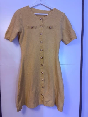 Beige stickad kortklänning med knappar - Supersöt beige stickad klänning med korta ärmar och dekorativa knappar hela vägen framtill. Klänningen har två små fickor vid bröstet med matchande knappar och ribbade detaljer vid ärmslut och nederkant. Perfekt för dig som gillar en enkel men chic look.