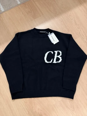 Svart stickad tröja Cole Buxton - Svart stickad tröja från Cole Buxton med vita CB-bokstäver på bröstet. Tröjan har rund hals och ribbade muddar vid ärmslut och nederkant. Tillverkad i 100% bomull, perfekt för dig som gillar stilrena och enkla plagg med en cool logga.