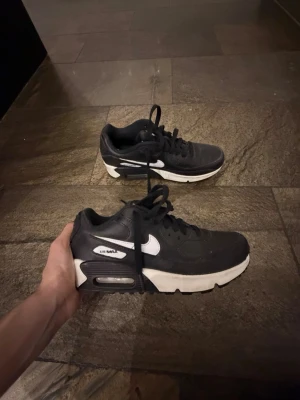Svarta Nike Air Max 90 sneakers - Säljer ett par klassiska svarta Nike Air Max 90 sneakers med vit sula och vit swoosh. Skorna har snörning och den ikoniska Air Max-detaljen i sulan. Perfekta för dig som gillar streetwear och vill ha en tidlös look.