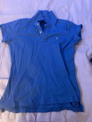 Blå pikétröja från Ralph Lauren storlek M - Klassisk blå pikétröja från Ralph Lauren med korta ärmar och vit knappslå framtill. Tröjan har den ikoniska lilla broderade loggan på bröstet och är tillverkad i mjuk bomull. Perfekt till jeans eller shorts för en avslappnad look.
