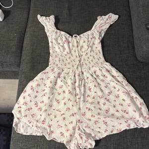 Vit blommig byxdress från FB Sister XS - Supersöt vit byxdress från FB Sister i storlek XS. Plagget har små rosa blommor, smockad midja och axelband med volang. Materialet är mjukt och lätt, perfekt för varma dagar. Knytning framtill ger extra detalj och passformen är avslappnad och luftig.